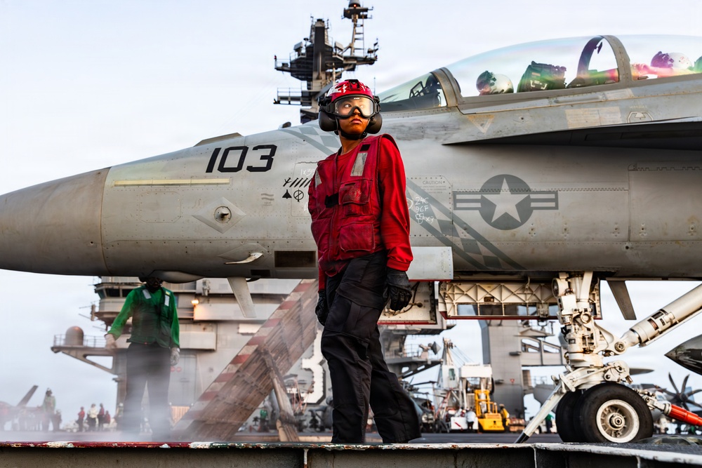 A U.S. Navy deck crew member in red jersey and safety gear stands on an aircraft carrier flight deck in front of a military jet marked '103'. The aircraft displays a U.S. military star insignia and appears to be an F/A-18 Hornet or Super Hornet. The scene captures active flight deck operations with multiple personnel and equipment visible in the background.