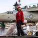 A U.S. Navy deck crew member in red jersey and safety gear stands on an aircraft carrier flight deck in front of a military jet marked '103'. The aircraft displays a U.S. military star insignia and appears to be an F/A-18 Hornet or Super Hornet. The scene captures active flight deck operations with multiple personnel and equipment visible in the background.