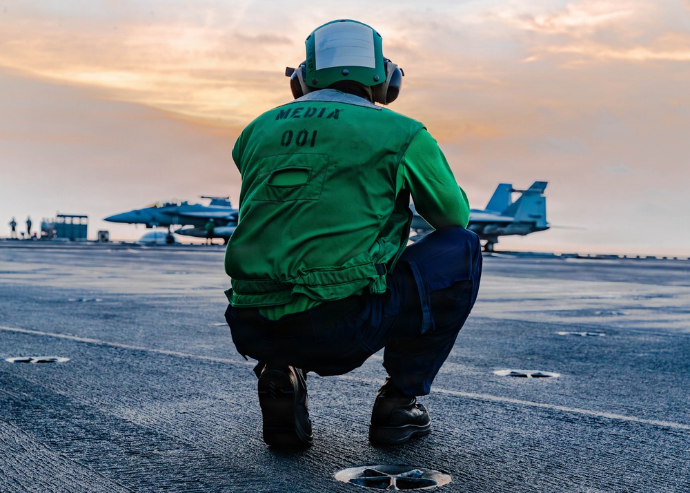 A person wearing a green vest labeled 'MEDIA 001' and a green flight helmet is crouching on the flight deck of an aircraft carrier at sunset or sunrise. Military jet aircraft, likely F/A-18 Hornets or Super Hornets, are visible in the background on the deck. The scene depicts a media personnel embedded with a naval carrier air wing during flight operations.
