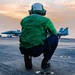 A person wearing a green vest labeled 'MEDIA 001' and a green flight helmet is crouching on the flight deck of an aircraft carrier at sunset or sunrise. Military jet aircraft, likely F/A-18 Hornets or Super Hornets, are visible in the background on the deck. The scene depicts a media personnel embedded with a naval carrier air wing during flight operations.
