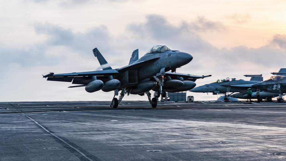 A carrier-based jet fighter, consistent with an F/A-18 Super Hornet, is captured at the moment of launch or landing on the flight deck of a naval aircraft carrier. The aircraft has its landing gear deployed and appears to be in motion, with additional aircraft visible in the background. The image is taken at dusk or dawn, with a warm-toned sky and overcast clouds providing dramatic lighting.