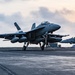 A carrier-based jet fighter, consistent with an F/A-18 Super Hornet, is captured at the moment of launch or landing on the flight deck of a naval aircraft carrier. The aircraft has its landing gear deployed and appears to be in motion, with additional aircraft visible in the background. The image is taken at dusk or dawn, with a warm-toned sky and overcast clouds providing dramatic lighting.