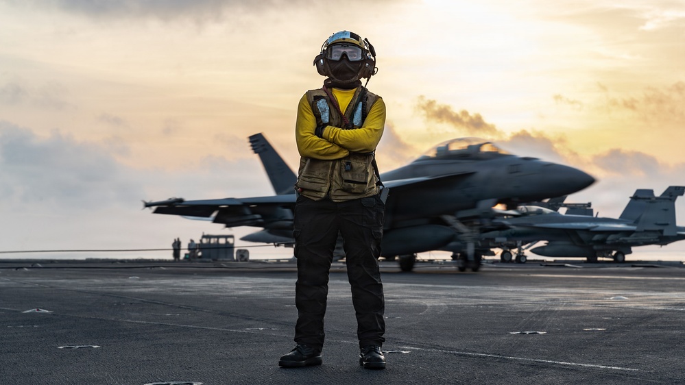 A naval aviation deck crew member stands on an aircraft carrier flight deck wearing a yellow jersey, tactical vest, helmet, and goggles with arms crossed. Multiple F/A-18 Hornet or Super Hornet jet aircraft are visible in the background on the flight deck. The image is taken during golden hour with a dramatic sunset sky backdrop.