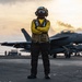 A naval aviation deck crew member stands on an aircraft carrier flight deck wearing a yellow jersey, tactical vest, helmet, and goggles with arms crossed. Multiple F/A-18 Hornet or Super Hornet jet aircraft are visible in the background on the flight deck. The image is taken during golden hour with a dramatic sunset sky backdrop.