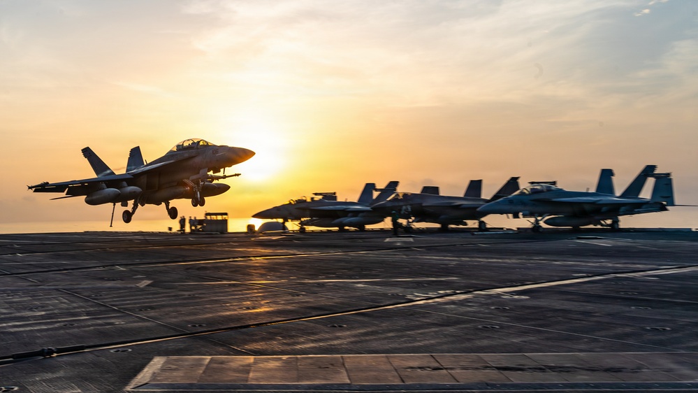A military jet aircraft is captured mid-launch or low-altitude flight above the deck of an aircraft carrier at sunset. Several additional fighter jets are parked in a row on the flight deck in the background. The golden sunset creates dramatic backlighting, silhouetting the aircraft against a warm orange and yellow sky.