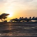 A military jet aircraft is captured mid-launch or low-altitude flight above the deck of an aircraft carrier at sunset. Several additional fighter jets are parked in a row on the flight deck in the background. The golden sunset creates dramatic backlighting, silhouetting the aircraft against a warm orange and yellow sky.