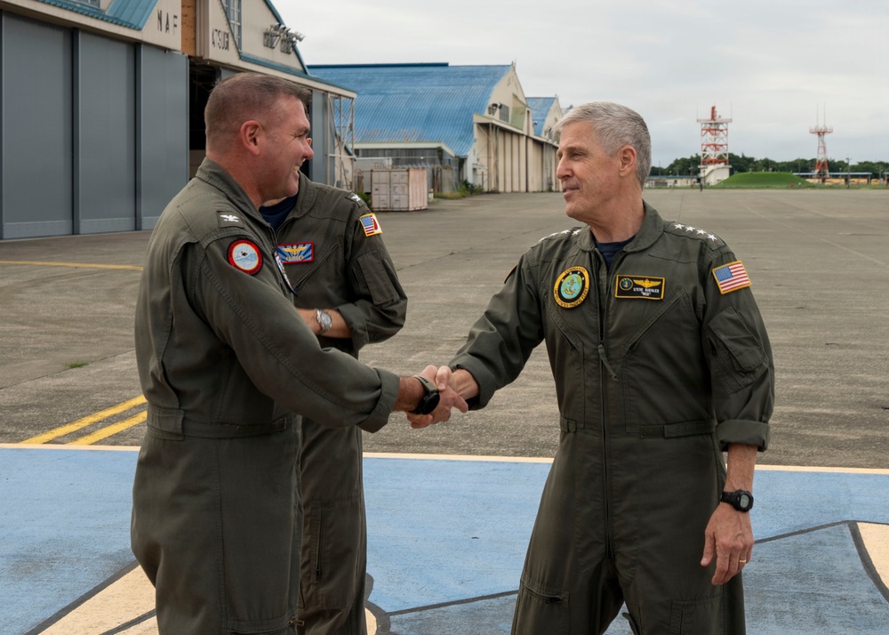 Two military personnel in olive green flight suits are shaking hands on an airfield tarmac. Both wear U.S. flag patches and unit insignia patches on their flight suits, with a third individual visible in the background. The setting appears to be a military air base with hangars visible in the background.