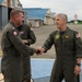 Two military personnel in olive green flight suits are shaking hands on an airfield tarmac. Both wear U.S. flag patches and unit insignia patches on their flight suits, with a third individual visible in the background. The setting appears to be a military air base with hangars visible in the background.