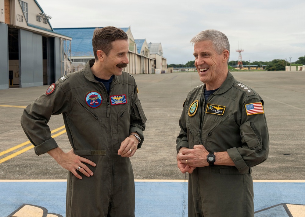 Two military personnel in olive green flight suits are conversing and smiling on an airfield tarmac. The individual on the right displays two stars on his shoulder indicating a Major General rank, along with an American flag patch and aviator wings. They appear to be engaged in a friendly, informal discussion in front of aircraft hangars.