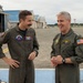 Two military personnel in olive green flight suits are conversing and smiling on an airfield tarmac. The individual on the right displays two stars on his shoulder indicating a Major General rank, along with an American flag patch and aviator wings. They appear to be engaged in a friendly, informal discussion in front of aircraft hangars.