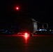 A large military transport aircraft, consistent with a C-17 Globemaster III, is photographed on a tarmac at night. The aircraft is illuminated primarily by its own red navigation/anti-collision lights, which cast a red glow on the ground beneath it. The dark background reveals distant airfield lighting along the horizon.