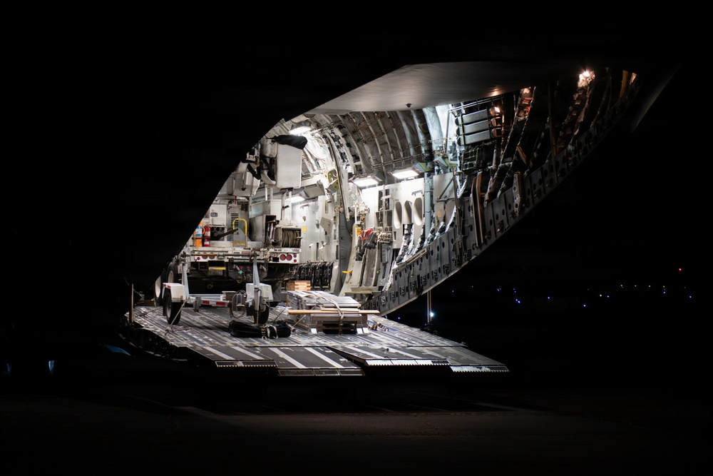 A large military or heavy-lift cargo aircraft is photographed at night with its rear cargo ramp fully lowered and open. The interior of the aircraft is illuminated, revealing the cargo bay with equipment and loading infrastructure visible. The surrounding environment is dark, consistent with a nighttime airfield or tarmac setting.