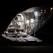 A large military or heavy-lift cargo aircraft is photographed at night with its rear cargo ramp fully lowered and open. The interior of the aircraft is illuminated, revealing the cargo bay with equipment and loading infrastructure visible. The surrounding environment is dark, consistent with a nighttime airfield or tarmac setting.