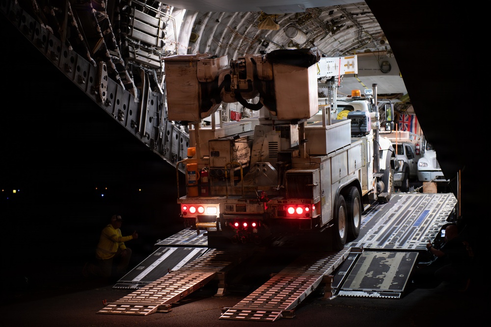 A large military or cargo aircraft is being loaded at night with heavy specialized vehicles via extended ramp systems. Multiple trucks and equipment are staged both inside and outside the aircraft, with ground crew visible overseeing the loading operation. A flag, appearing to be the Union Jack (British flag), is partially visible in the background near the aircraft entrance.