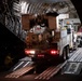 A large military or cargo aircraft is being loaded at night with heavy specialized vehicles via extended ramp systems. Multiple trucks and equipment are staged both inside and outside the aircraft, with ground crew visible overseeing the loading operation. A flag, appearing to be the Union Jack (British flag), is partially visible in the background near the aircraft entrance.