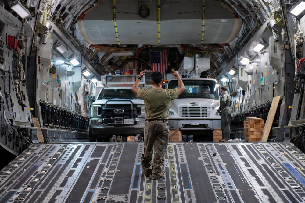 A military loadmaster is directing vehicles onto a large military cargo aircraft, likely a C-17 Globemaster III, using hand signals. Two vehicles — a Ford F-series pickup truck and a Freightliner medium-duty truck — are being loaded into the aircraft's cargo bay. An American flag is visible in the background near the front of the cargo hold.