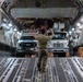 A military loadmaster is directing vehicles onto a large military cargo aircraft, likely a C-17 Globemaster III, using hand signals. Two vehicles — a Ford F-series pickup truck and a Freightliner medium-duty truck — are being loaded into the aircraft's cargo bay. An American flag is visible in the background near the front of the cargo hold.