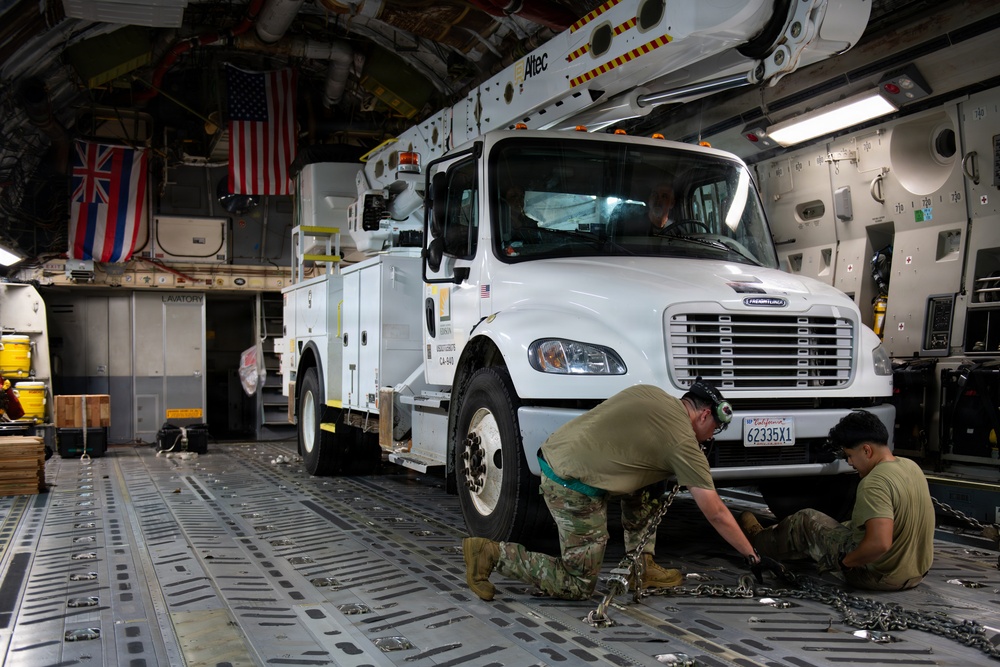 Military personnel are securing a white Freightliner utility truck with an Altec aerial lift inside the cargo hold of a large military transport aircraft, likely a C-17 Globemaster. Two soldiers in camouflage uniforms are working at the front of the vehicle, attaching chains to tie-down points on the aircraft floor. Flags visible in the background include the United States flag and what appears to be the Hawaii state flag, suggesting this may be a relief or support mission to Hawaii.