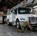 Military personnel are securing a white Freightliner utility truck with an Altec aerial lift inside the cargo hold of a large military transport aircraft, likely a C-17 Globemaster. Two soldiers in camouflage uniforms are working at the front of the vehicle, attaching chains to tie-down points on the aircraft floor. Flags visible in the background include the United States flag and what appears to be the Hawaii state flag, suggesting this may be a relief or support mission to Hawaii.