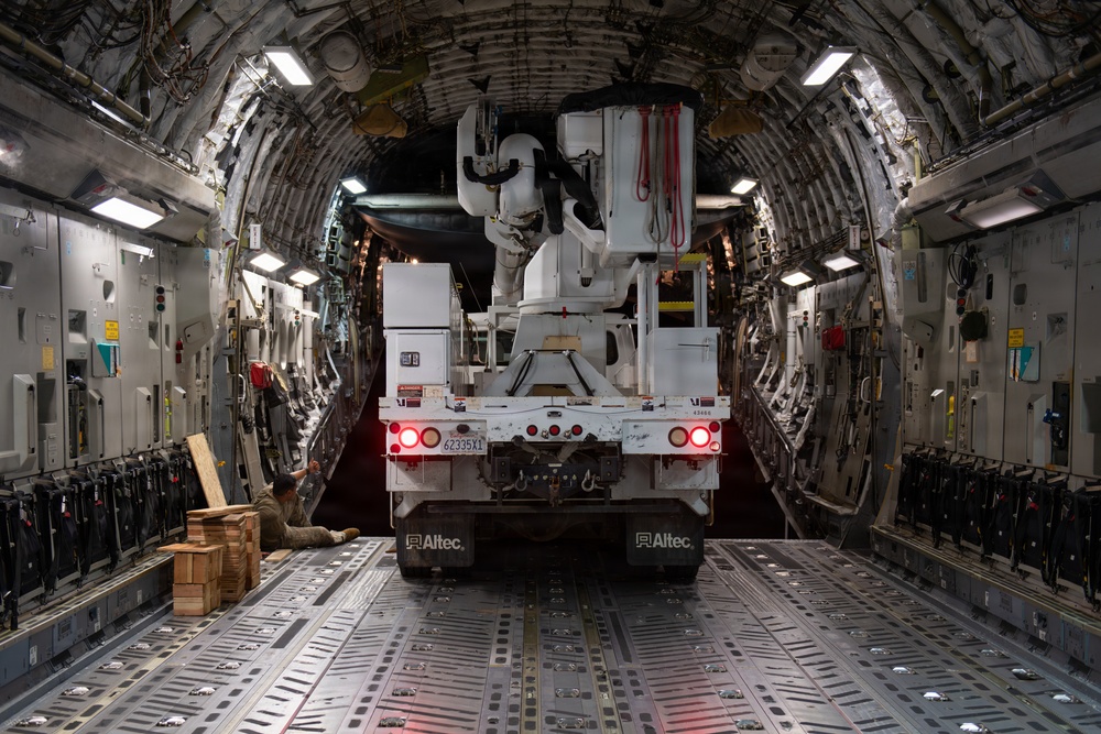The interior of a large military cargo aircraft, likely a C-17 Globemaster III, showing a white Altec utility/bucket truck being loaded or secured inside the cargo hold. A person is visible on the left side of the aircraft, seated on the floor near stacked wooden blocks, apparently involved in the securing process. The vehicle's brake lights are illuminated, and the aircraft's ribbed fuselage interior with tie-down rails is clearly visible.