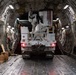 The interior of a large military cargo aircraft, likely a C-17 Globemaster III, showing a white Altec utility/bucket truck being loaded or secured inside the cargo hold. A person is visible on the left side of the aircraft, seated on the floor near stacked wooden blocks, apparently involved in the securing process. The vehicle's brake lights are illuminated, and the aircraft's ribbed fuselage interior with tie-down rails is clearly visible.