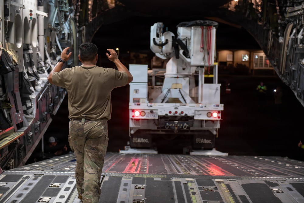 A military loadmaster is directing a large utility truck (branded 'Altec') as it backs into the cargo hold of what appears to be a C-17 Globemaster III military transport aircraft. The scene is taking place at night on an airfield tarmac. The personnel is using hand signals to guide the vehicle safely onto the aircraft's cargo ramp.