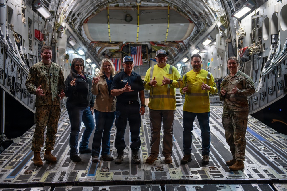 Seven individuals are posing inside the cargo hold of a large military transport aircraft, likely a C-17 Globemaster III. The group includes two military personnel in uniform (one Navy, one Army/Air Force in MultiCam), civilian workers including one in a high-visibility safety vest, and others in casual attire. All individuals are making 'shaka' (hang loose) hand gestures toward the camera, suggesting a celebratory or commemorative photo.