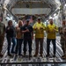Seven individuals are posing inside the cargo hold of a large military transport aircraft, likely a C-17 Globemaster III. The group includes two military personnel in uniform (one Navy, one Army/Air Force in MultiCam), civilian workers including one in a high-visibility safety vest, and others in casual attire. All individuals are making 'shaka' (hang loose) hand gestures toward the camera, suggesting a celebratory or commemorative photo.