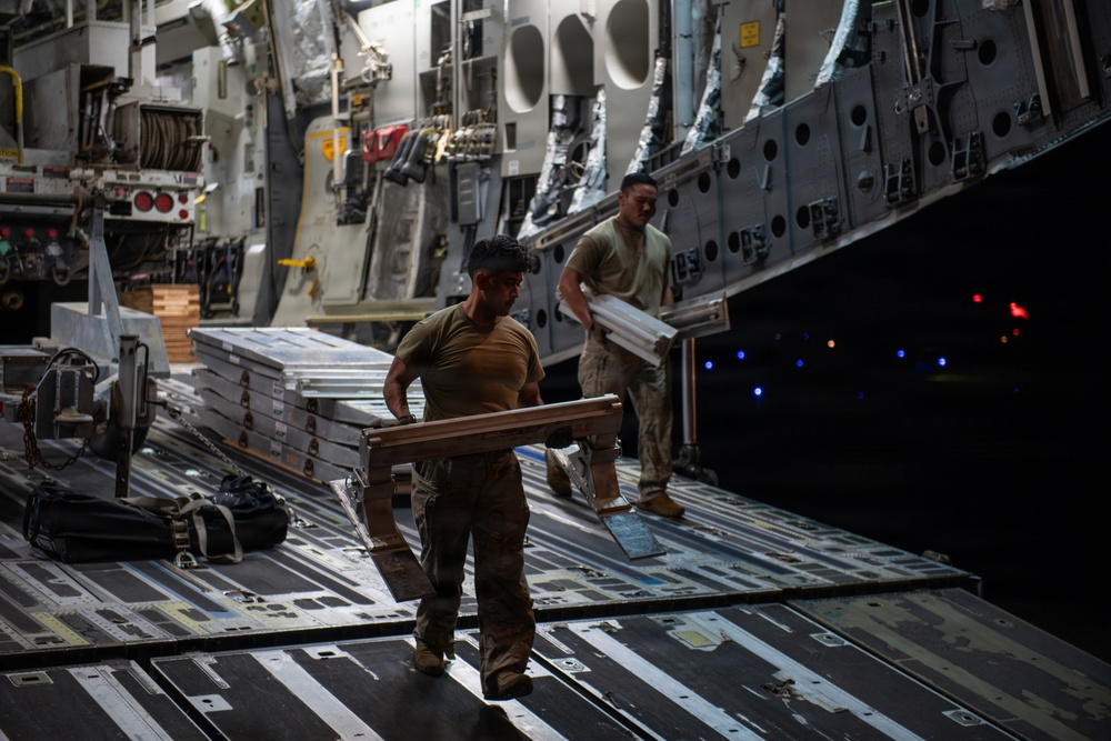 Two military personnel in combat uniforms are loading or unloading cargo inside what appears to be a large military transport aircraft, likely a C-17 Globemaster III. They are handling flat, aluminum-framed equipment cases or panels on the aircraft's cargo floor. The interior lighting is dim, with the cargo ramp area illuminated by ambient and indicator lights.