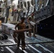 Two military personnel in combat uniforms are loading or unloading cargo inside what appears to be a large military transport aircraft, likely a C-17 Globemaster III. They are handling flat, aluminum-framed equipment cases or panels on the aircraft's cargo floor. The interior lighting is dim, with the cargo ramp area illuminated by ambient and indicator lights.