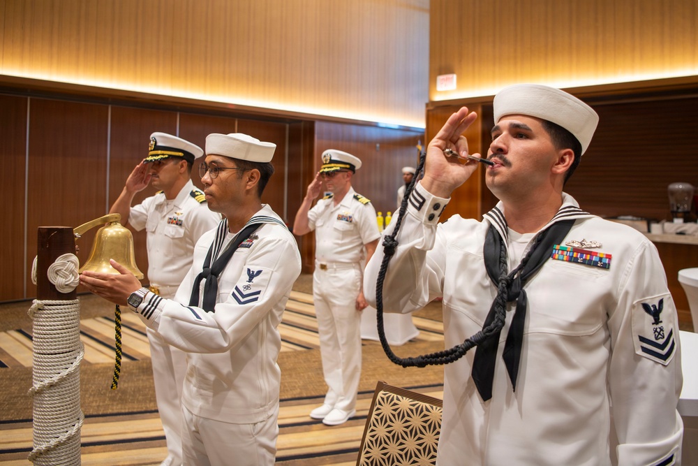 A formal U.S. Navy ceremonial event taking place in what appears to be a hotel ballroom or conference facility. Enlisted sailors in dress white uniforms are performing a side-boy ceremony, with one sailor piping (using a boatswain's pipe) and another ringing a ship's bell, while officers in the background render salutes. The scene depicts a traditional naval honors ceremony, likely welcoming a dignitary or senior officer.