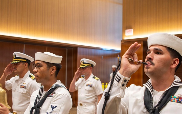 A formal U.S. Navy ceremonial event taking place in what appears to be a hotel ballroom or conference facility. Enlisted sailors in dress white uniforms are performing a side-boy ceremony, with one sailor piping (using a boatswain's pipe) and another ringing a ship's bell, while officers in the background render salutes. The scene depicts a traditional naval honors ceremony, likely welcoming a dignitary or senior officer.