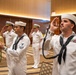 A formal U.S. Navy ceremonial event taking place in what appears to be a hotel ballroom or conference facility. Enlisted sailors in dress white uniforms are performing a side-boy ceremony, with one sailor piping (using a boatswain's pipe) and another ringing a ship's bell, while officers in the background render salutes. The scene depicts a traditional naval honors ceremony, likely welcoming a dignitary or senior officer.
