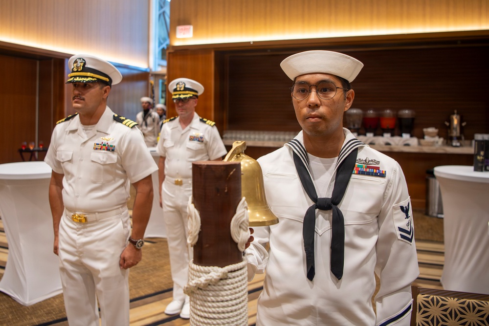 A formal U.S. Navy ceremony is taking place in what appears to be a hotel ballroom or conference facility. A junior enlisted sailor in dress whites stands at attention beside a brass ship's bell mounted on a wooden post with rope coiling. Senior officers in dress white uniforms with gold insignia and ribbons stand in the background.