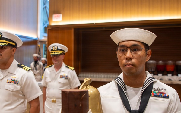 A formal U.S. Navy ceremony is taking place in what appears to be a hotel ballroom or conference facility. A junior enlisted sailor in dress whites stands at attention beside a brass ship's bell mounted on a wooden post with rope coiling. Senior officers in dress white uniforms with gold insignia and ribbons stand in the background.