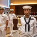 A formal U.S. Navy ceremony is taking place in what appears to be a hotel ballroom or conference facility. A junior enlisted sailor in dress whites stands at attention beside a brass ship's bell mounted on a wooden post with rope coiling. Senior officers in dress white uniforms with gold insignia and ribbons stand in the background.