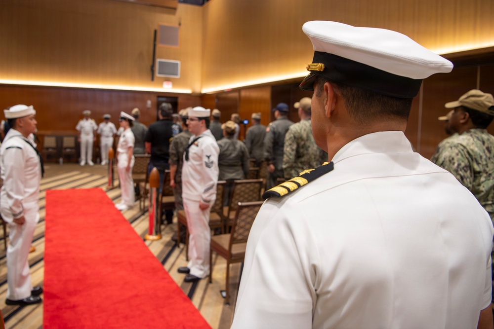 A formal U.S. Navy military ceremony is taking place in an indoor venue with wood-paneled walls. A red carpet runner is laid down the center aisle, flanked by personnel in various uniforms including white dress uniforms and camouflage utilities. The foreground features a senior officer in white dress uniform with gold sleeve stripes, viewed from behind.