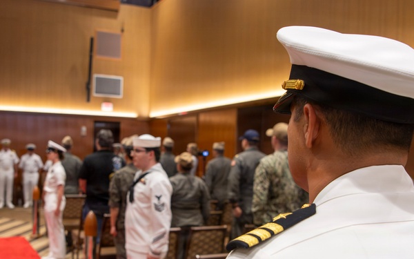 A formal U.S. Navy military ceremony is taking place in an indoor venue with wood-paneled walls. A red carpet runner is laid down the center aisle, flanked by personnel in various uniforms including white dress uniforms and camouflage utilities. The foreground features a senior officer in white dress uniform with gold sleeve stripes, viewed from behind.