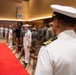 A formal U.S. Navy military ceremony is taking place in an indoor venue with wood-paneled walls. A red carpet runner is laid down the center aisle, flanked by personnel in various uniforms including white dress uniforms and camouflage utilities. The foreground features a senior officer in white dress uniform with gold sleeve stripes, viewed from behind.