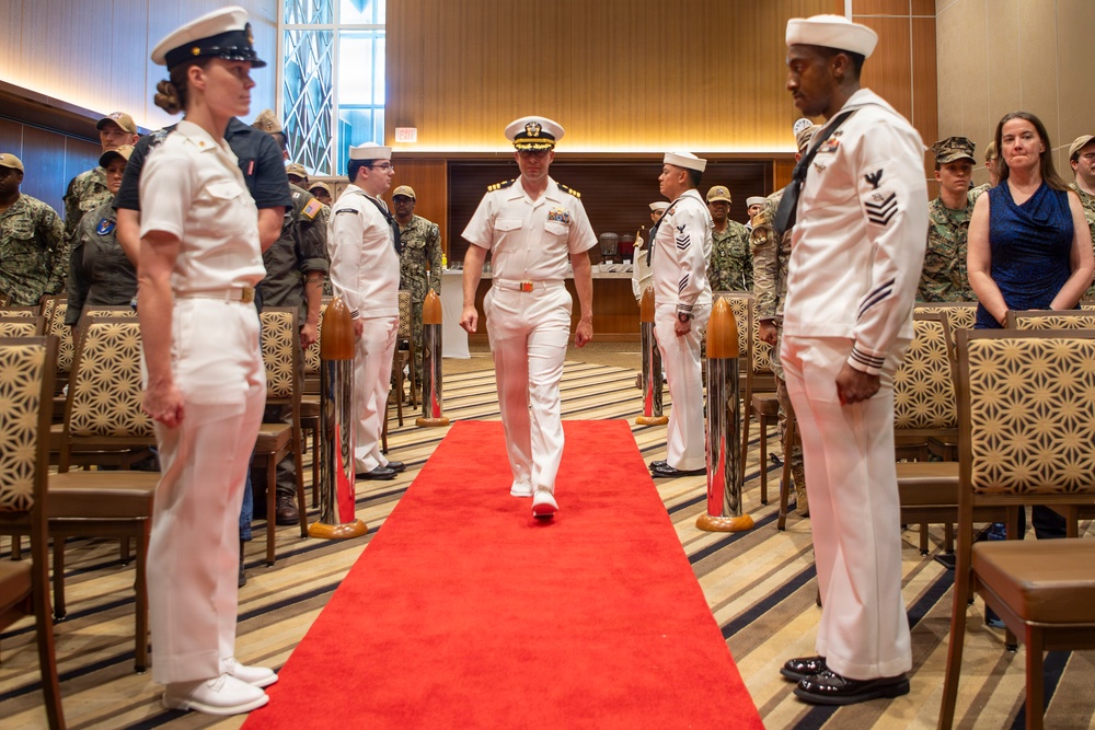 A formal U.S. Navy military ceremony is taking place in an indoor venue with a red carpet aisle. A senior naval officer in dress white uniform walks down the center aisle flanked by personnel in various uniforms standing at attention. The setting appears to be a change of command or retirement ceremony based on the formal arrangement and mixed military attendance.
