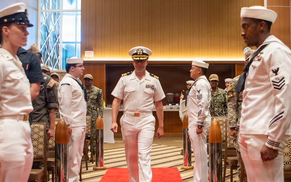 A formal U.S. Navy military ceremony is taking place in an indoor venue with a red carpet aisle. A senior naval officer in dress white uniform walks down the center aisle flanked by personnel in various uniforms standing at attention. The setting appears to be a change of command or retirement ceremony based on the formal arrangement and mixed military attendance.