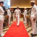 A formal U.S. Navy military ceremony is taking place in an indoor venue with a red carpet aisle. A senior naval officer in dress white uniform walks down the center aisle flanked by personnel in various uniforms standing at attention. The setting appears to be a change of command or retirement ceremony based on the formal arrangement and mixed military attendance.