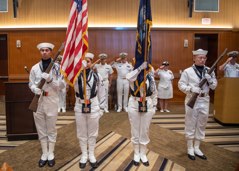 A U.S. Navy color guard ceremony is taking place in an indoor venue, likely a conference or banquet hall. Four sailors in dress white uniforms with white gloves and covers are presenting the American flag and a Navy flag while carrying ceremonial rifles. Behind them, officers and enlisted personnel in various white dress uniforms stand at attention or render salutes.