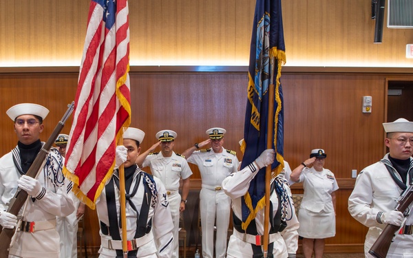 A U.S. Navy color guard ceremony is taking place in an indoor venue, likely a conference or banquet hall. Four sailors in dress white uniforms with white gloves and covers are presenting the American flag and a Navy flag while carrying ceremonial rifles. Behind them, officers and enlisted personnel in various white dress uniforms stand at attention or render salutes.