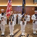 A U.S. Navy color guard ceremony is taking place in an indoor venue, likely a conference or banquet hall. Four sailors in dress white uniforms with white gloves and covers are presenting the American flag and a Navy flag while carrying ceremonial rifles. Behind them, officers and enlisted personnel in various white dress uniforms stand at attention or render salutes.