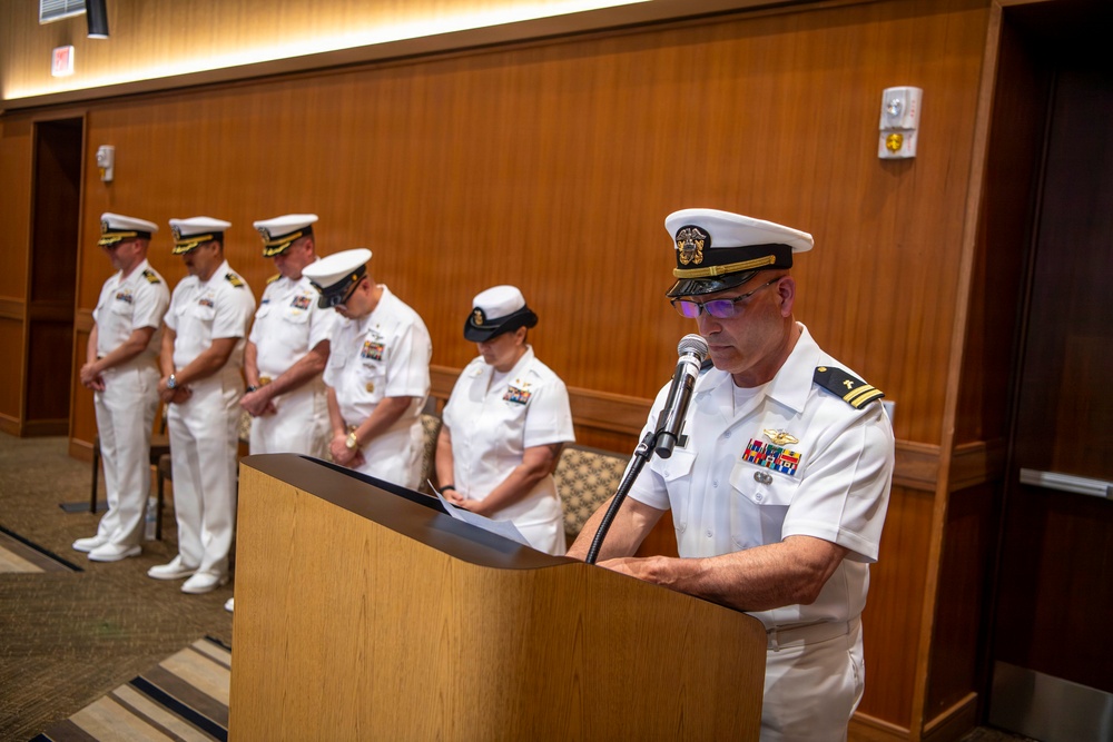 A U.S. Navy officer in white dress uniform stands at a wooden podium speaking into a microphone, appearing to conduct or lead a formal military ceremony. Behind him, a line of five Navy personnel in white dress uniforms stand at attention or with heads bowed, suggesting a prayer, invocation, or solemn moment. The setting appears to be an indoor conference or ceremonial room with wood-paneled walls.