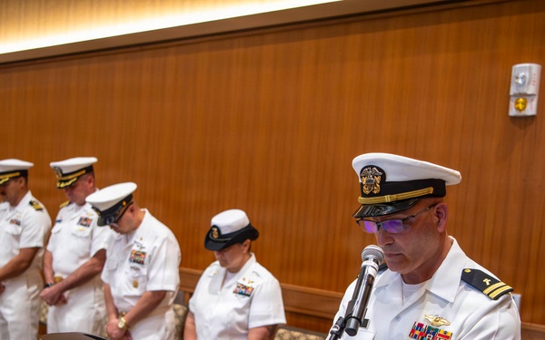 A U.S. Navy officer in white dress uniform stands at a wooden podium speaking into a microphone, appearing to conduct or lead a formal military ceremony. Behind him, a line of five Navy personnel in white dress uniforms stand at attention or with heads bowed, suggesting a prayer, invocation, or solemn moment. The setting appears to be an indoor conference or ceremonial room with wood-paneled walls.