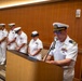 A U.S. Navy officer in white dress uniform stands at a wooden podium speaking into a microphone, appearing to conduct or lead a formal military ceremony. Behind him, a line of five Navy personnel in white dress uniforms stand at attention or with heads bowed, suggesting a prayer, invocation, or solemn moment. The setting appears to be an indoor conference or ceremonial room with wood-paneled walls.