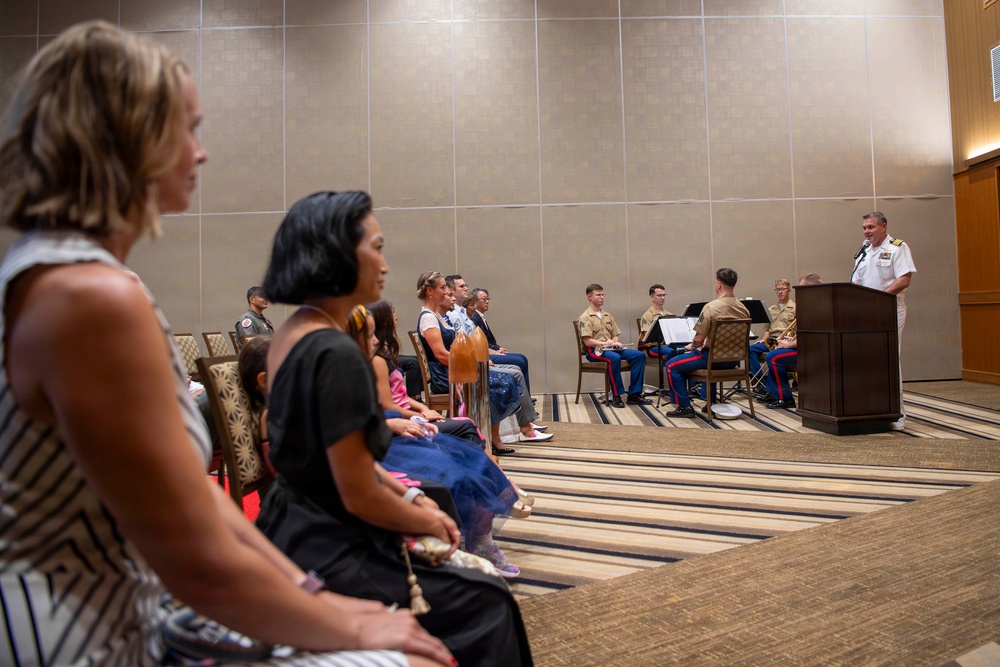 A formal military ceremony is taking place in a hotel ballroom or conference room. A naval officer in white dress uniform is speaking at a podium, while an audience of civilians and uniformed Marines with musical instruments are seated facing him. The event appears to be an official military function such as a promotion, retirement, or award ceremony.