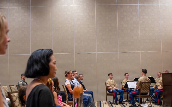 A formal military ceremony is taking place in a hotel ballroom or conference room. A naval officer in white dress uniform is speaking at a podium, while an audience of civilians and uniformed Marines with musical instruments are seated facing him. The event appears to be an official military function such as a promotion, retirement, or award ceremony.